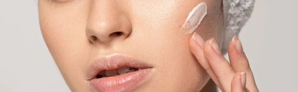 Foto panoramica di giovane donna che applica crema cosmetica sul viso, isolata su grigio — Foto stock