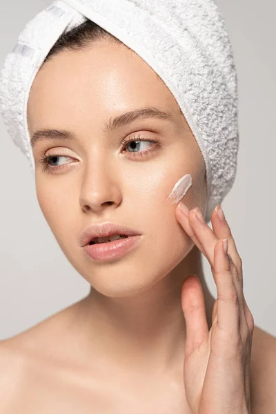 Jolie femme avec serviette sur la tête appliquer crème hydratante sur le visage, isolé sur gris — Photo de stock