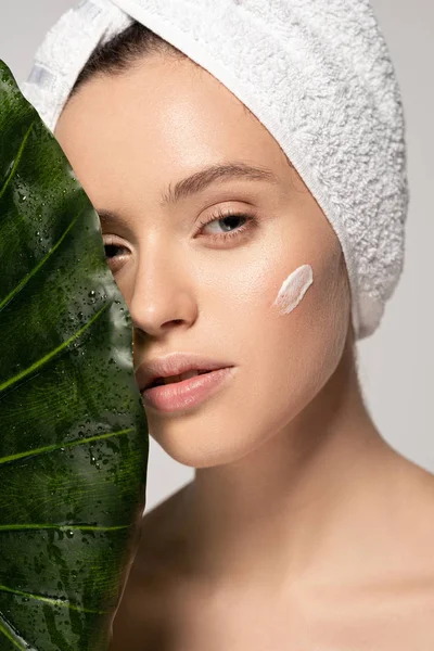 Menina bonita com creme cosmético no rosto e toalha na cabeça posando com folha verde, isolado em cinza — Fotografia de Stock