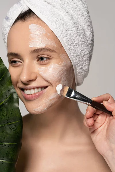 Happy woman applying mask on face with cosmetic brush, isolated on grey with leaf — Stock Photo