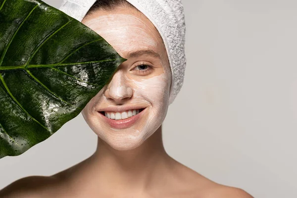 Щаслива молода жінка з косметичною маскою на обличчі та рушником на голові позує зеленим листом, ізольована на сірому — стокове фото