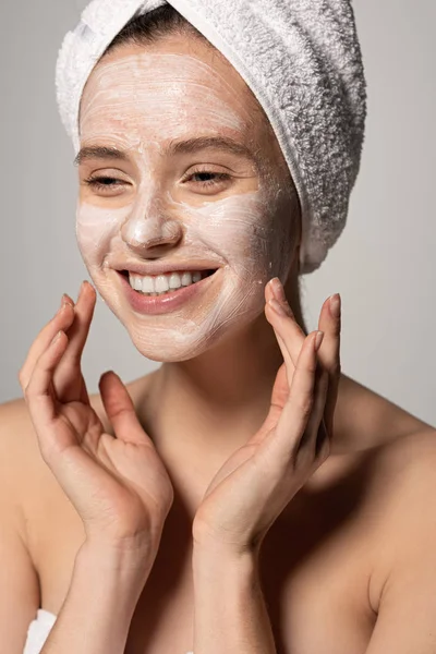 Menina feliz atraente com máscara cosmética no rosto e toalha na cabeça, isolado em cinza — Fotografia de Stock