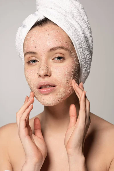 Jolie jeune femme avec serviette sur la tête appliquant gommage de noix de coco sur le visage, isolé sur gris — Photo de stock