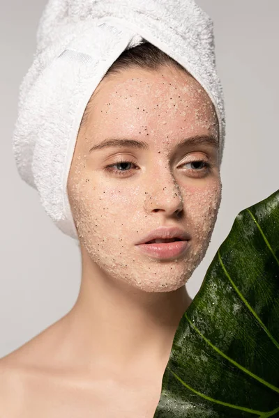 Attraktives Mädchen mit Handtuch auf dem Kopf und Kokosnuss-Peeling im Gesicht, das mit Blatt posiert, isoliert auf grau — Stockfoto