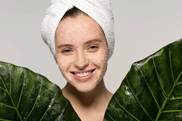 Lächelndes Mädchen mit Handtuch auf dem Kopf und Kokospeeling im Gesicht, das mit Blättern posiert, isoliert auf grau — Stockfoto