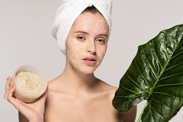 Menina atraente com toalha na cabeça segurando folha verde e recipiente de plástico com esfoliação de coco, isolado em cinza — Fotografia de Stock