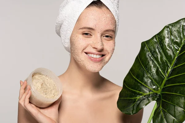 Smiling girl with towel on head holding green leaf and plastic container with coconut scrub, isolated on grey — Stock Photo