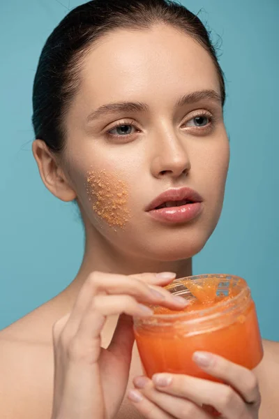 Hermosa chica aplicando amarillo azúcar exfoliante de plástico contenedor, aislado en azul - foto de stock