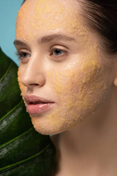 Menina atraente com esfoliação de açúcar no rosto segurando folha, isolado em azul — Fotografia de Stock