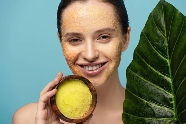 Hermosa chica alegre sosteniendo tazón de madera con exfoliante de azúcar, aislado en azul - foto de stock