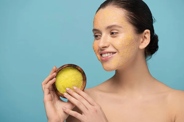 Mujer joven feliz sosteniendo un tazón de madera con exfoliante de azúcar, aislado en azul - foto de stock