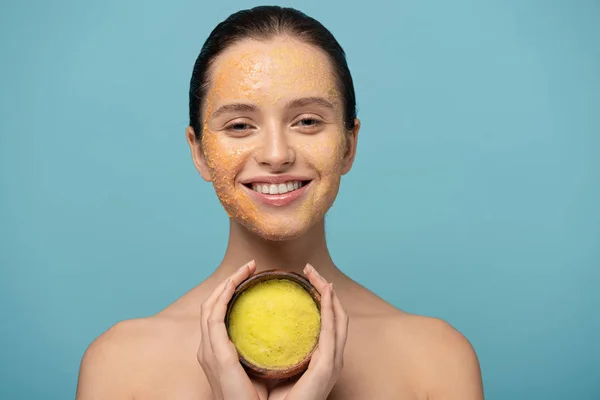 Cheerful girl holding wooden bowl with sugar scrub, isolated on blue — Stock Photo