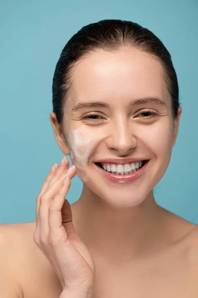 Sorrindo menina aplicando espuma de limpeza no rosto, isolado em azul — Fotografia de Stock