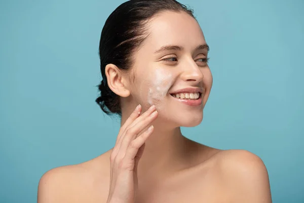 Atractiva chica sonriente aplicando espuma limpiadora en la cara, aislado en azul - foto de stock