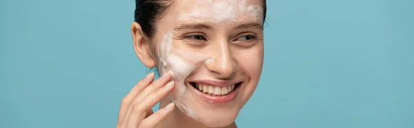 Colpo panoramico di allegra giovane donna che applica schiuma detergente sul viso, isolata sul blu — Foto stock