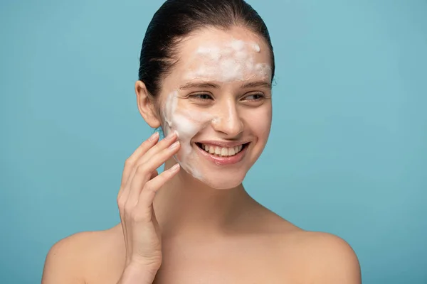 Bela jovem feliz aplicando espuma de limpeza no rosto, isolado em azul — Fotografia de Stock