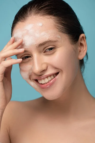 Schönes lächelndes Mädchen, das Reinigungsschaum auf das Gesicht aufträgt, isoliert auf blau — Stockfoto