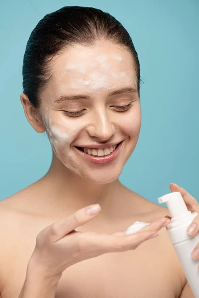 Fille souriante appliquer de la mousse nettoyante de bouteille sur le visage, isolé sur le bleu — Photo de stock