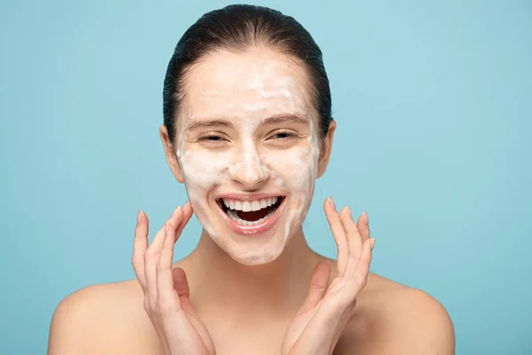 Attraente ragazza eccitata che applica schiuma detergente sul viso, isolata sul blu — Foto stock