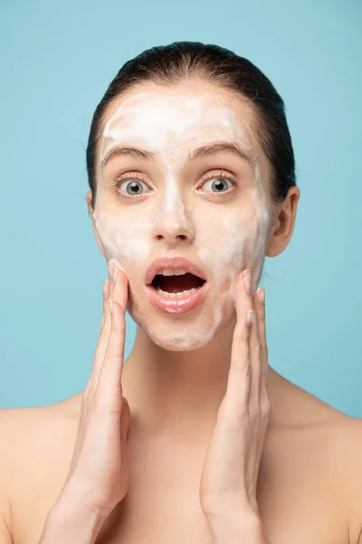 Chica impactada aplicando espuma limpiadora en la cara, aislado en azul - foto de stock