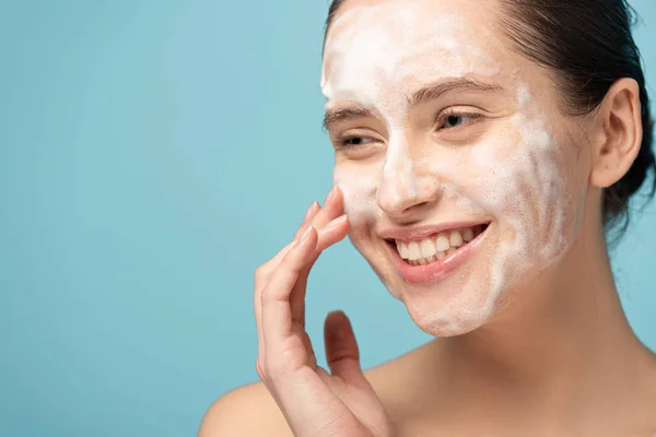Bela mulher sorridente aplicando espuma de limpeza no rosto, isolado em azul — Fotografia de Stock