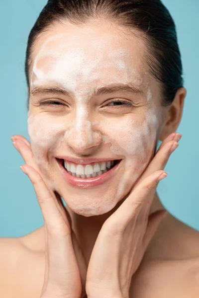 Bela sorrindo jovem mulher aplicando espuma de limpeza no rosto, isolado em azul — Fotografia de Stock