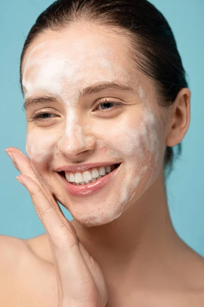 Attraktives glückliches Mädchen, das Reinigungsschaum auf das Gesicht aufträgt, isoliert auf blau — Stockfoto