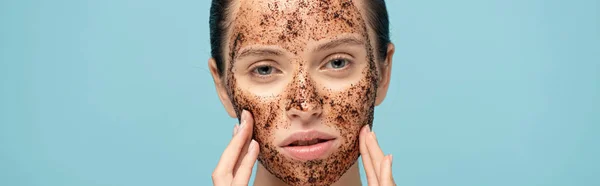 Colpo panoramico di ragazza attraente applicare scrub caffè sul viso, isolato su blu — Foto stock