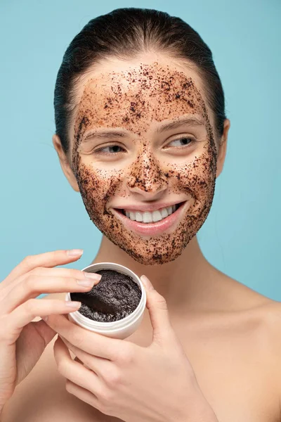 Fille souriante appliquant gommage de café de récipient en plastique, isolé sur bleu — Photo de stock