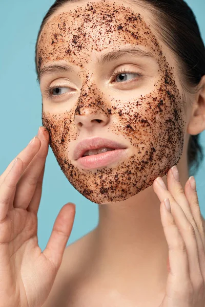 Bella ragazza tenera applicando scrub caffè sul viso, isolato su blu — Foto stock