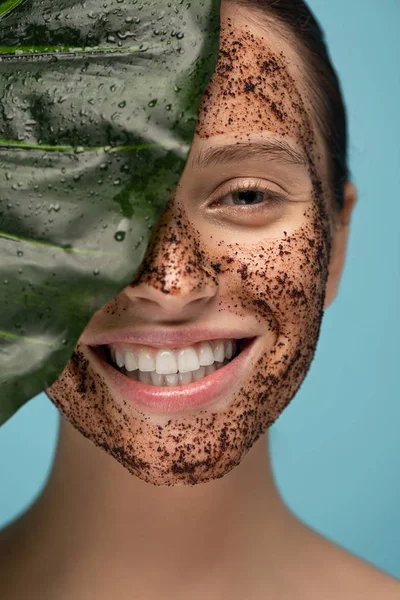 Beautiful happy girl with coffee scrub on face, isolated on blue with leaf — Stock Photo