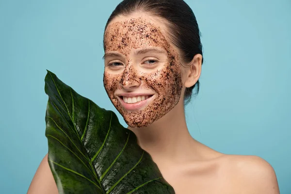Beautiful cheerful woman with coffee scrub on face holding leaf, isolated on blue — Stock Photo