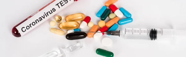 Панорамный снимок красочных таблеток рядом с образцом с коронавирусной надписью и шприцем на белом — стоковое фото