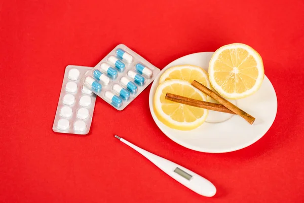 Sliced lemons on saucer with cinnamon sticks near pills and digital thermometer on red — Stock Photo