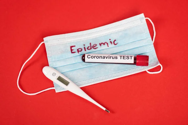 Вид зверху зразка з тестом на коронавірус поблизу захисної медичної маски з епідемічним написанням та цифровим термометром на червоному — стокове фото