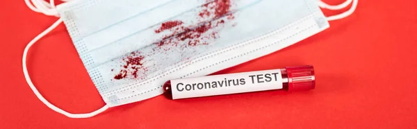 Prise de vue panoramique de l'échantillon avec coronavirus test lettrage près du masque médical avec du sang sur rouge — Photo de stock