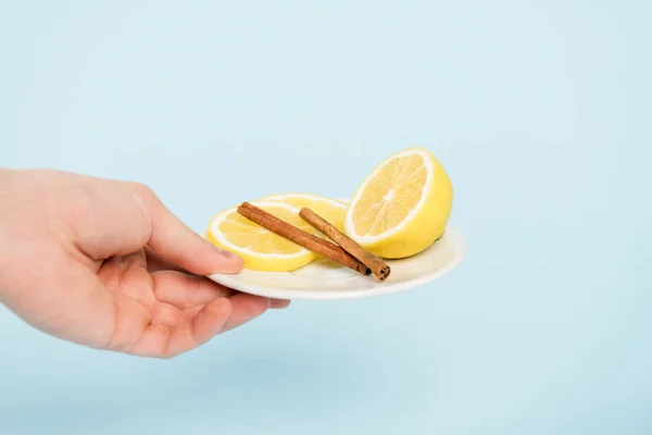 Cropped view of man holding plate with sliced lemons and cinnamon sticks isolated on blue — Stock Photo