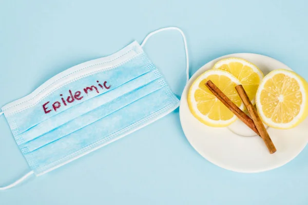 Untertasse mit Zimtstangen und Zitronen in der Nähe medizinischer Maske mit Epidemie-Schriftzug auf blau — Stockfoto