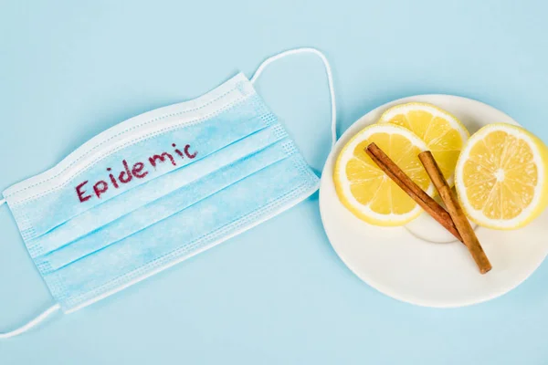 Vista superior del platillo con palitos de canela y limones cerca de la máscara médica con letras epidémicas en azul - foto de stock