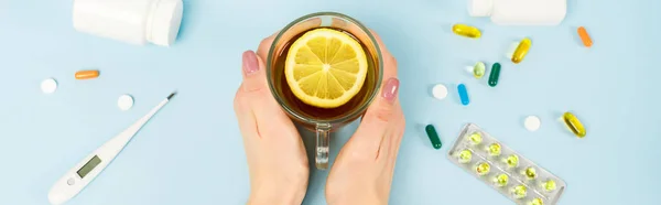 Plan panoramique de femme tenant une tasse de thé avec du citron tranché près de pilules et thermomètre numérique sur bleu — Photo de stock