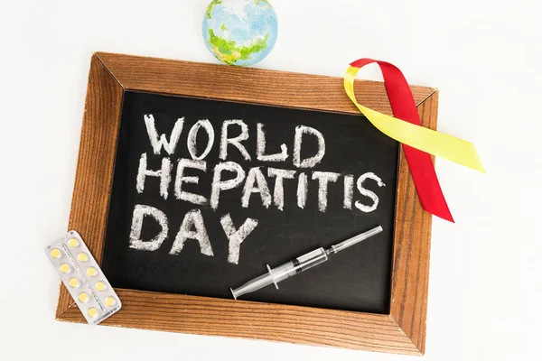 Top view of chalkboard with world hepatitis day lettering near ribbon, globe, pills and syringe isolated on white — Stock Photo