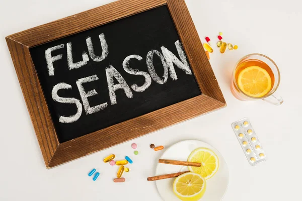 Top view of chalkboard with flu season lettering near pills and tea with lemon on white — Stock Photo