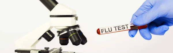 Panoramic shot of scientist in latex glove holding test tube with flu test lettering near microscope isolated on white — Stock Photo