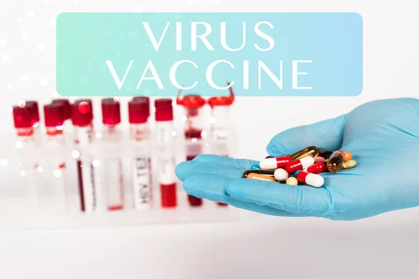 Vista recortada del científico en guantes de látex sosteniendo pastillas cerca de tubos de ensayo y letras de vacunas contra virus en blanco - foto de stock