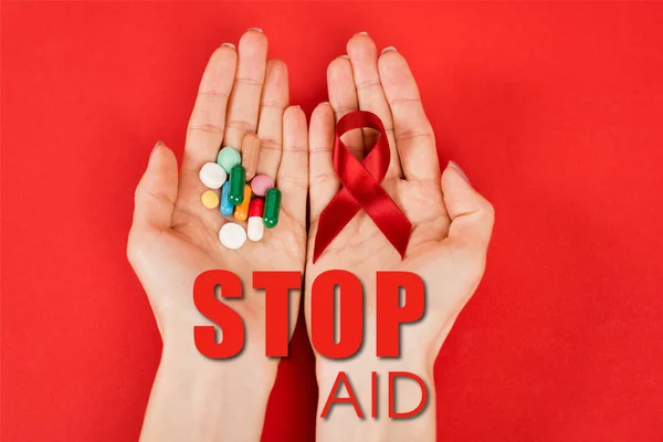 Vue du dessus de la femme tenant un ruban rouge et des pilules près de l'arrêt lettrage de l'aide sur rouge — Photo de stock