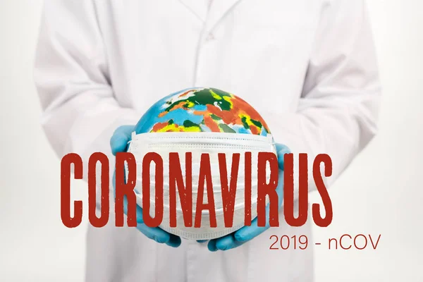 Cropped view of scientist holding globe in protective mask near coronavirus, 2019-ncov lettering on white — Stock Photo