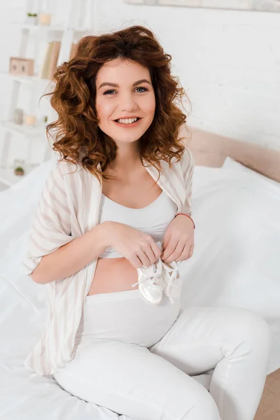 Belle femme enceinte tenant des chaussons de bébé et souriant à la caméra sur le lit — Photo de stock