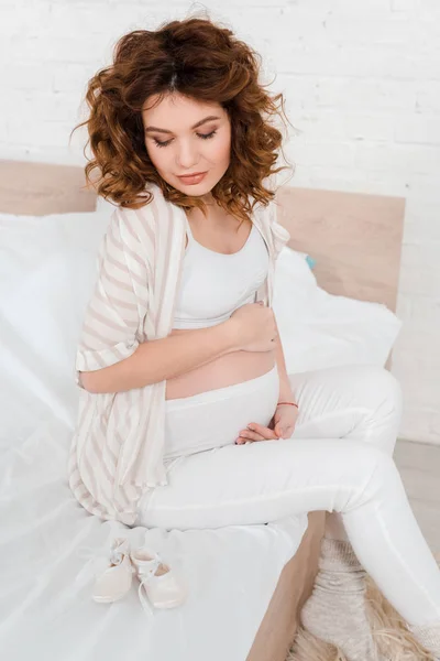 Hermosa mujer embarazada tocando vientre cerca de botines de bebé en la cama - foto de stock