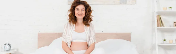 Schöne schwangere Frau lächelt in die Kamera, während sie auf dem Bett sitzt, Panoramaaufnahme — Stockfoto