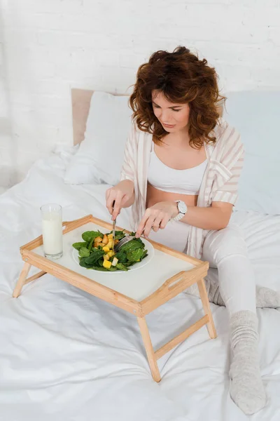 Vista de ángulo alto de la mujer embarazada comiendo ensalada cerca del vaso de leche en la bandeja del desayuno en la cama - foto de stock